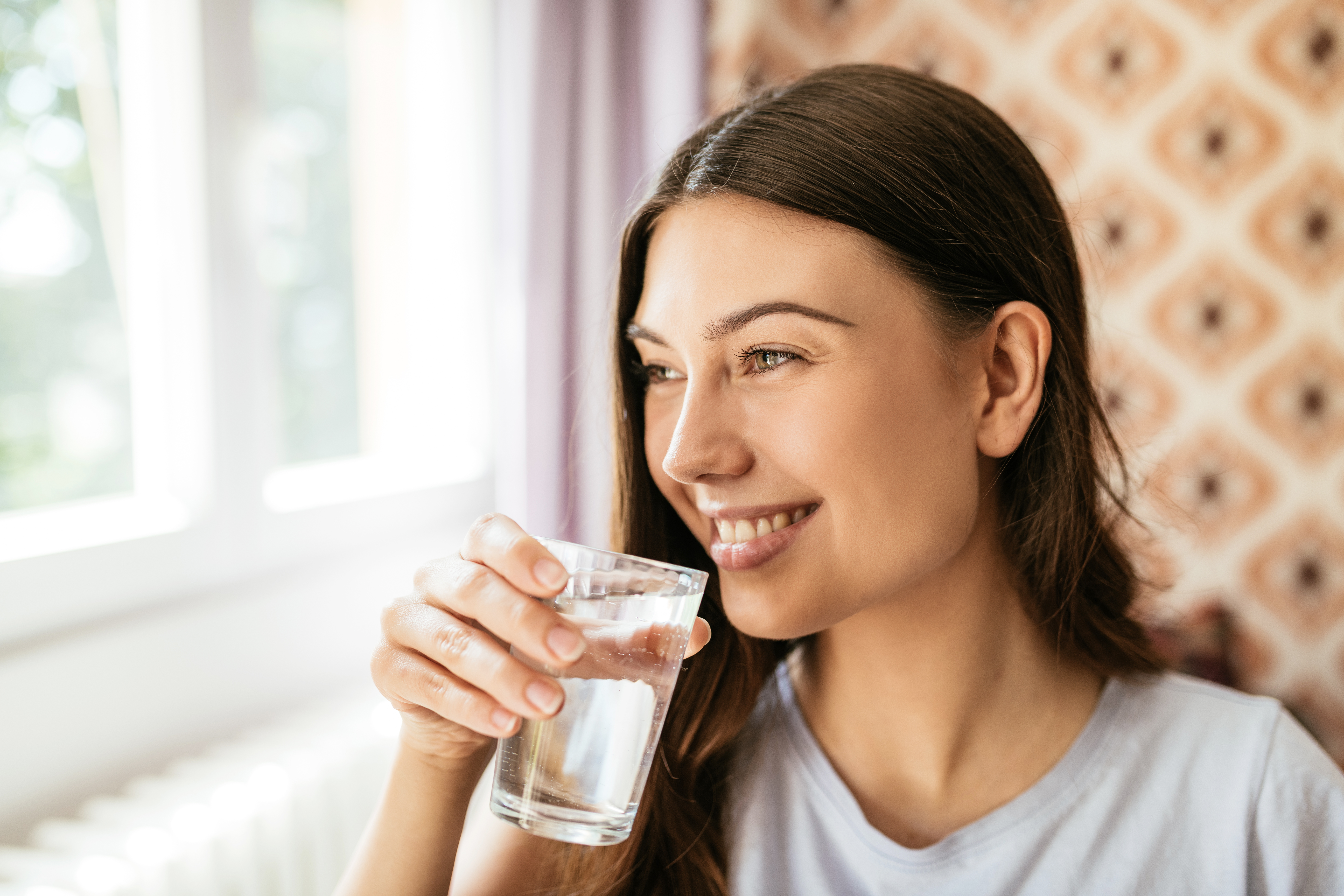 Healthy young woman drink water after taking a birth control pill while looking out the window and smiling.