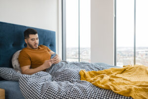A man wearing pajamas in his apartment bedroom on a sunny winters morning. He is lying down and using his smartphone as he relaxes in bed.