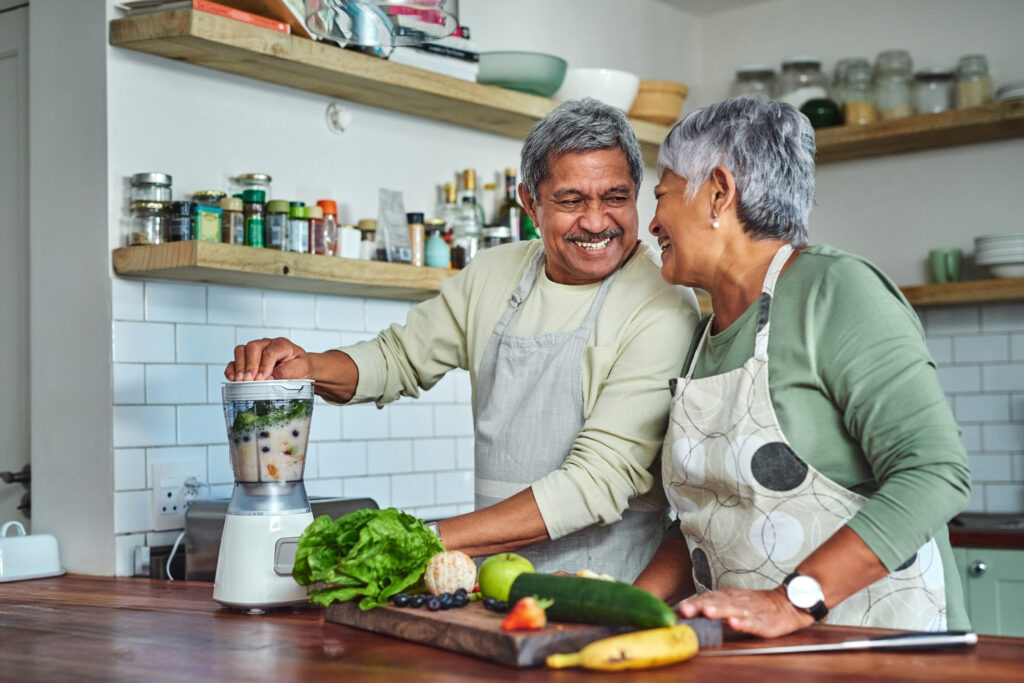 A happy couple laughs while making a smoothie in their kitchen, surrounded by fresh fruits and vegetables.