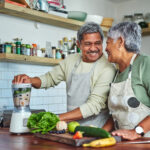 A happy couple laughs while making a smoothie in their kitchen, surrounded by fresh fruits and vegetables.