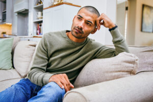A young man sitting on his couch with his hand on his head, reflecting on the personal impact of common misconceptions about herpes.