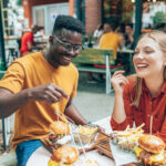 A young man and woman enjoying a meal of burgers and fries at an outdoor café, representing common dietary choices that may impact cholesterol levels.