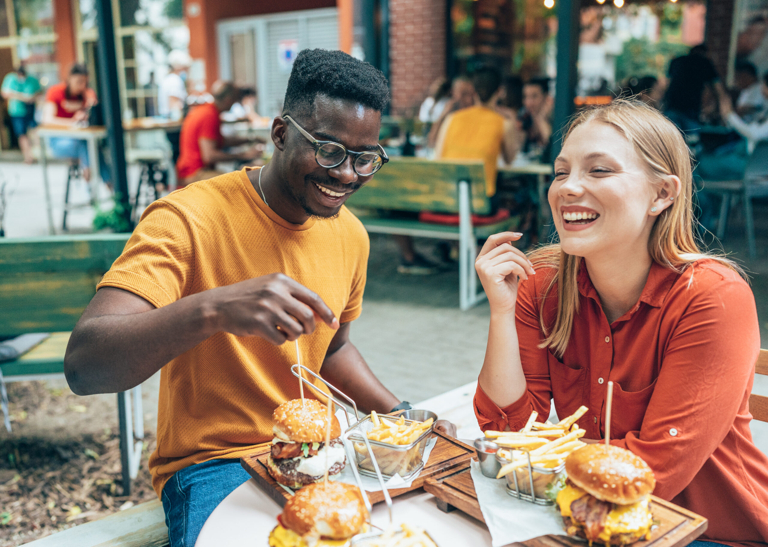 A young man and woman enjoying a meal of burgers and fries at an outdoor café, representing common dietary choices that may impact cholesterol levels.