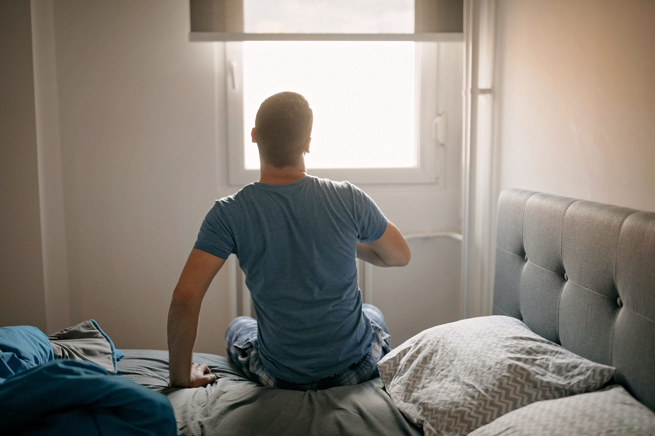 Man sitting on the edge of a bed, facing a bright window, reflecting on personal health decisions after a herpes outbreak.