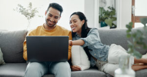 A couple sitting on their couch, smiling while using a laptop to research Viagra® and its uses.