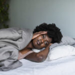 A young woman lying in bed in pain, holding her head, illustrating the struggles and misconceptions associated with insomnia.