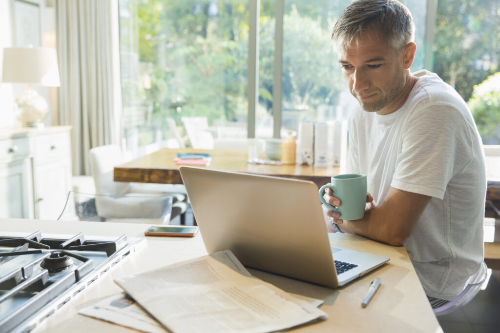 A man drinking coffee and working on his laptop in a kitchen, symbolizing a focus on personal well-being and productivity, reflecting the considerations of Stendra®.