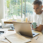 A man drinking coffee and working on his laptop in a kitchen, symbolizing a focus on personal well-being and productivity, reflecting the considerations of Stendra®.