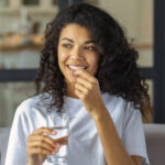 A woman smiling and holding an escitalopram (Lexapro®) pill and a glass of water, taking her medication to manage depression and anxiety.