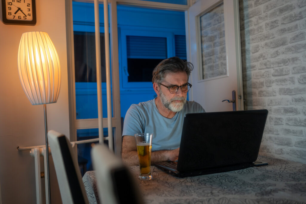 A man sitting at home at a table, drinking a beer while researching on his laptop to learn more about whether Cialis® is a suitable option for his health needs.