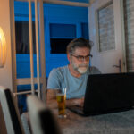 A man sitting at home at a table, drinking a beer while researching on his laptop to learn more about whether Cialis® is a suitable option for his health needs.