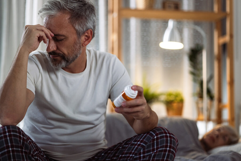 Man sitting on the edge of his bed, looking tired and frustrated, reading the bottle of Zoloft® (sertraline) in his hand, which may help address the emotional challenges or side effects he is experiencing