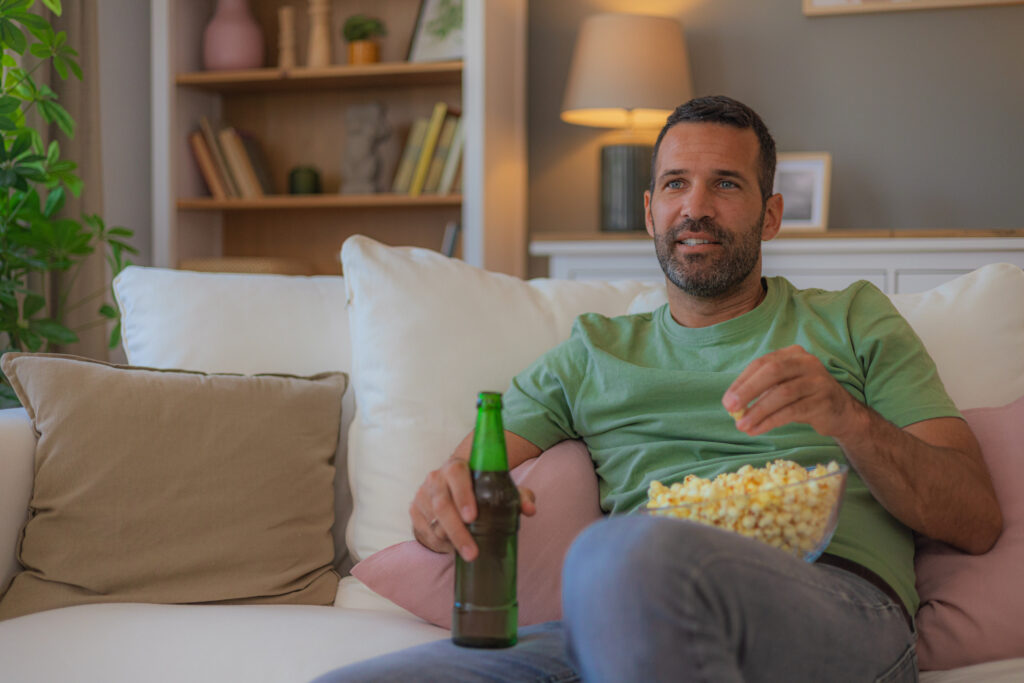 Man sitting on the couch watching TV with a beer and popcorn, unaware of the potential effects of alcohol consumption while taking finasteride for hair loss and prostate health.