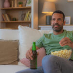 Man sitting on the couch watching TV with a beer and popcorn, unaware of the potential effects of alcohol consumption while taking finasteride for hair loss and prostate health.