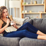 A woman sitting on the couch with a spoon, eating chocolate directly from the jar, symbolizing the comfort of emotional eating and cravings triggered by stress or sadness.