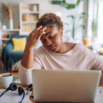 Woman feeling stressed at home, holding her head in worry while working on a laptop, symbolizing the mental health challenges that can impact weight loss success.