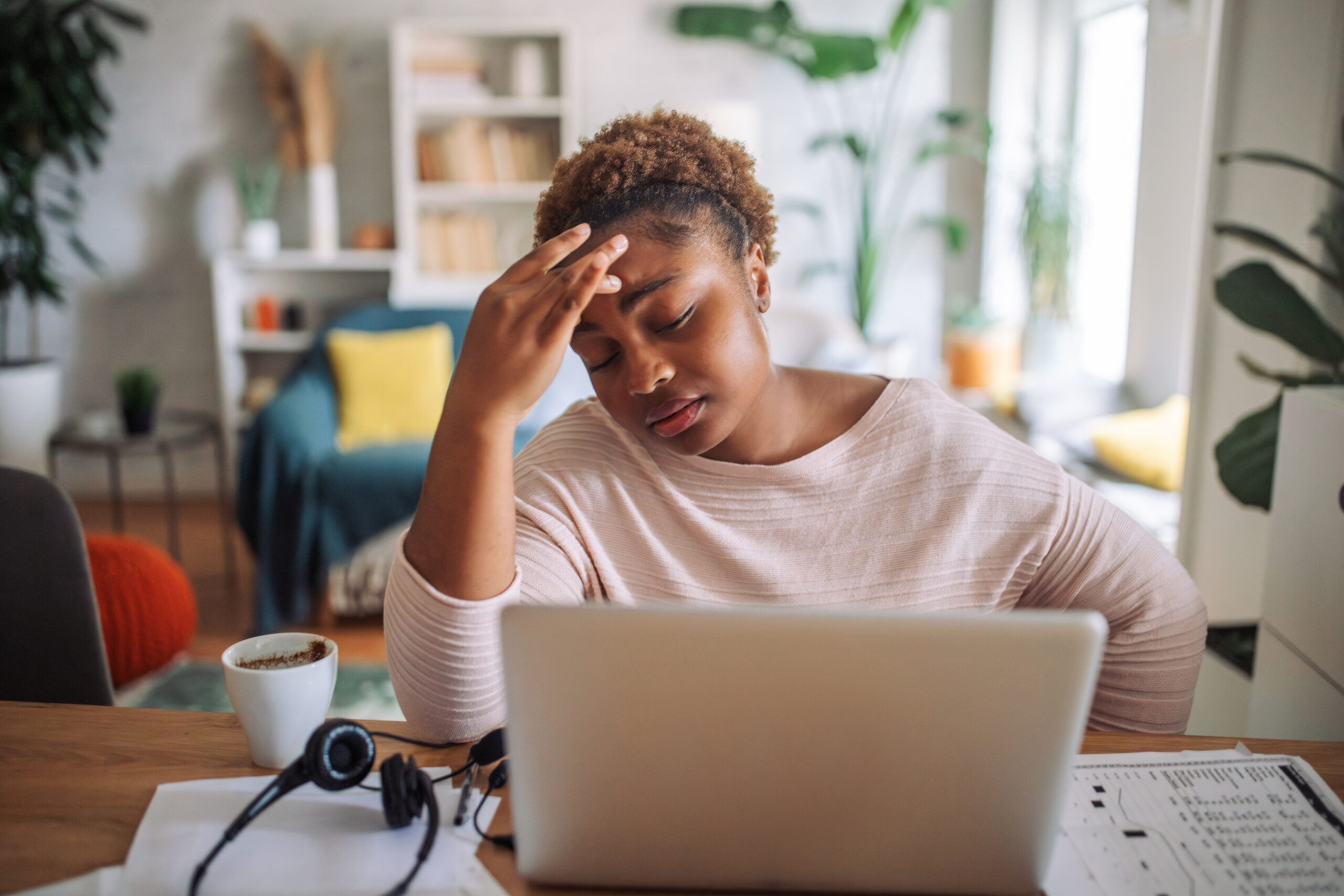 Woman feeling stressed at home, holding her head in worry while working on a laptop, symbolizing the mental health challenges that can impact weight loss success.