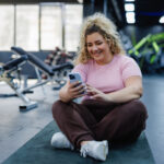 Woman checking her phone while on a gym mat after finishing her workout, highlighting the importance of exercise as part of her weight loss plan alongside her prescribed microdosing of semaglutide.