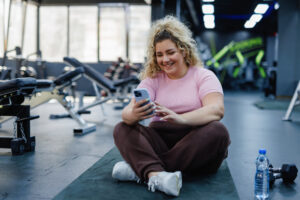 Woman checking her phone while on a gym mat after finishing her workout, highlighting the importance of exercise as part of her weight loss plan alongside her prescribed microdosing of semaglutide.