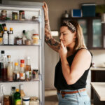 A woman standing in front of an open refrigerator, considering what to eat for dinner, symbolizing mindful eating and recognizing hunger signals to make healthier food choices.