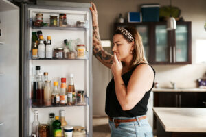 A woman standing in front of an open refrigerator, considering what to eat for dinner, symbolizing mindful eating and recognizing hunger signals to make healthier food choices.