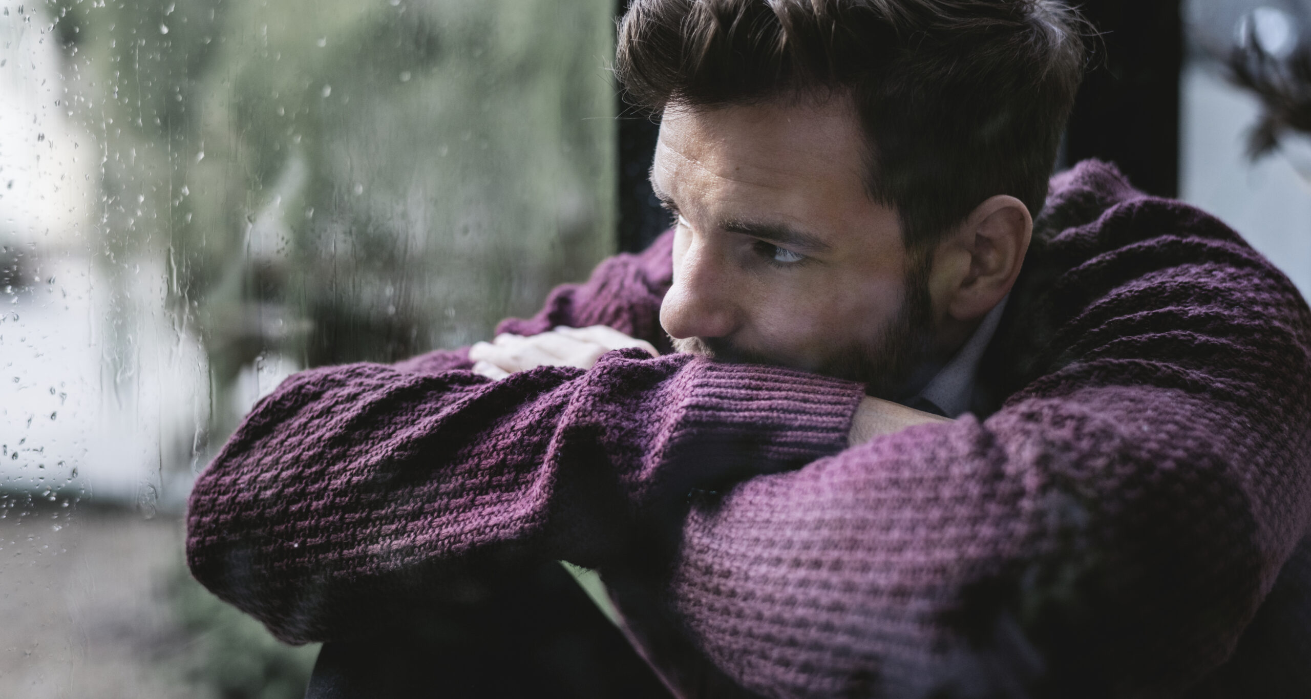 Sad and tired man looking out the window on a dark, rainy day, reflecting the emotional impact of seasonal affective disorder (SAD).