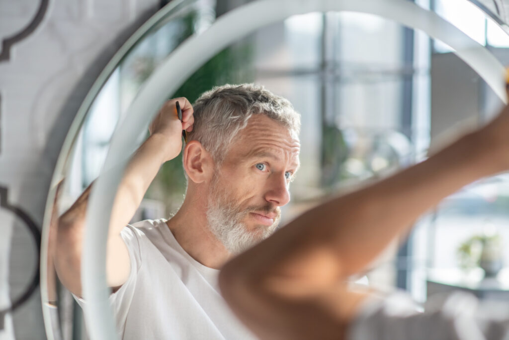 Man combing his hair while looking in the mirror, noticing thinning hair and hoping finasteride can help reduce symptoms of male pattern baldness.