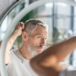 Man combing his hair while looking in the mirror, noticing thinning hair and hoping finasteride can help reduce symptoms of male pattern baldness.