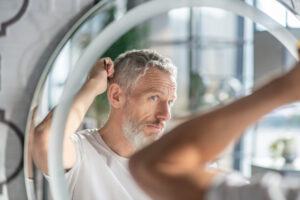 Man combing his hair while looking in the mirror, noticing thinning hair and hoping finasteride can help reduce symptoms of male pattern baldness.